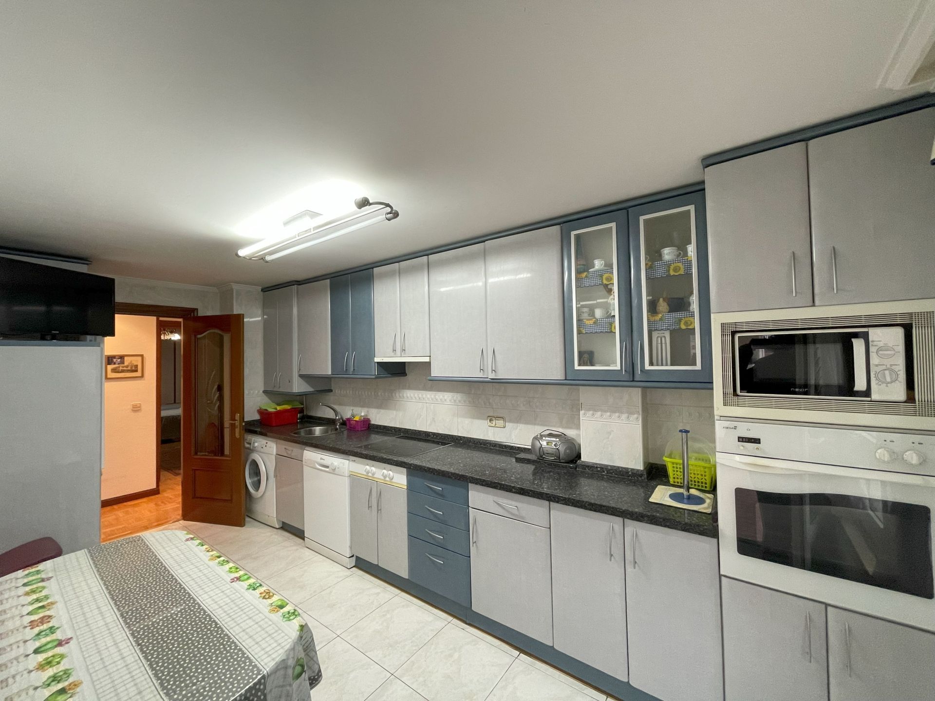 Cocina con muebles de madera gris combinados con encimera de piedra blanca  y armarios de pequeños electrodomésticos vitrocerámica y horno microondas  junto a la puerta de entrada a un apartamento de alquiler
